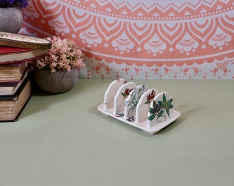 May include: White ceramic toast rack with a floral design featuring green leaves and red flowers. The rack has four slots for holding toast.