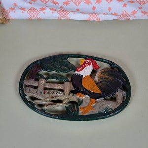 May include: A green cast iron hot pad featuring a rooster design. The rooster is painted in shades of red, white, yellow, and black. The rooster is standing on a fence with a green bush behind it.