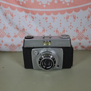 May include: A silver and black 35mm film camera with a black leatherette covering. The camera has a lens with a silver ring and a black shutter button. The camera has a silver top plate with the word "Sprint" printed on it.