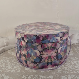 May include: A circular hat box featuring a floral design in purple, pink, and blue hues. It has a matching lid and a lavender rope handle. This decorative box is ideal for storage.