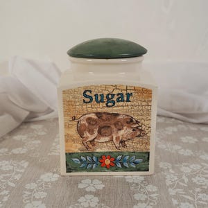 May include: A cream-coloured ceramic sugar container with a green lid. The container has a pig illustration and the word "Sugar". The design includes a cracked background and floral accents.