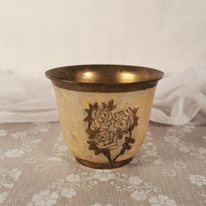 Pode incluir: Um vaso decorativo em estilo vintage com aro e base em latão. O exterior é creme com um desenho floral em castanho escuro. O vaso é provavelmente feito de metal e é adequado para uso interno.