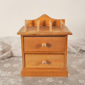 May include: A light brown wooden storage chest featuring two horizontal drawers with round knobs. The top surface of the chest includes a raised back panel and several vertical wooden pegs arranged in rows for organizing small items.