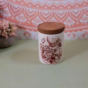 May include: A white ceramic jar with a brown wooden lid. The jar has a red and white illustration of a kitchen scene with a clock, a stove, and a teapot.