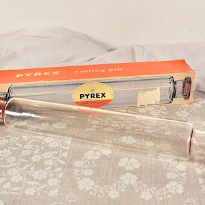 Può includere: Un mattarello Pyrex trasparente con tappi rossi, esposto con la sua scatola arancione originale. La scatola è etichettata "rolling pin" e presenta il logo Pyrex. Il mattarello è in vetro.