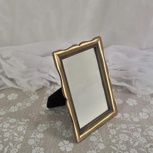 May include: A gold-coloured, rectangular picture frame with a scalloped edge. The frame has a dark interior border and a black velvet easel back. The frame is empty, ready to display a photograph or artwork.