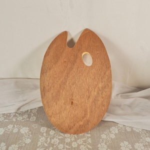 May include: An oval-shaped wooden artist palette with a natural light brown wood grain finish. The palette features a smooth surface, a thumb hole positioned near the top right, and a distinct cutout notch at the top centre for holding brushes.