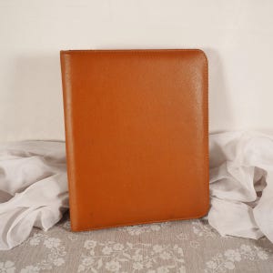 以下が含まれることがあります： 滑らかで均一な質感と丸みを帯びた角を持つ、長方形のタンカラーのレザー製ポートフォリオまたはドキュメントフォルダー。アイテムは閉じられており、端に沿ってきれいな直線状のステッチが施され、全体的にしっかりとした均一な外観を呈しています。