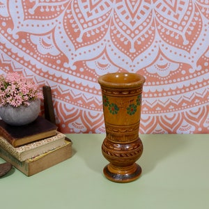 Puede incluir: Un jarrón de madera con acabado marrón claro, con detalles tallados y acentos florales verdes. El jarrón tiene una parte superior ancha y una base bulbosa, de aproximadamente 15 cm de altura. El fondo incluye un tapiz floral y una pequeña planta en maceta.