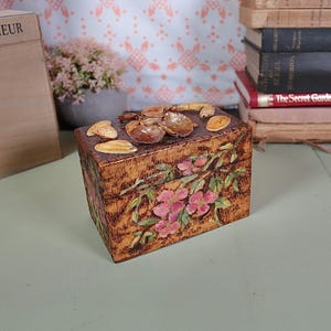 May include: A small wooden box with a floral design painted on the lid. The box is decorated with seashells and has a rustic, vintage look.
