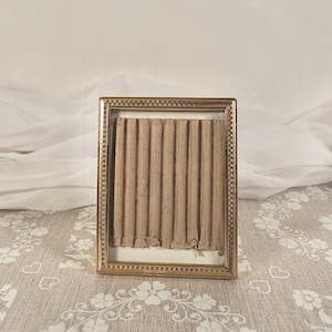 Pode incluir: Uma moldura retangular dourada com uma borda decorativa. A moldura contém um pedaço de papelão ondulado. O fundo apresenta um tecido branco e uma toalha de mesa estampada.