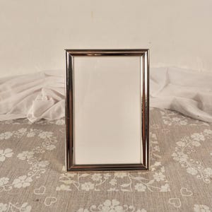 May include: A rectangular picture frame with a polished silver-tone metallic finish and a clean, simple border design. The frame is oriented vertically and features a plain white insert, providing a neutral display area for a photograph or artwork.