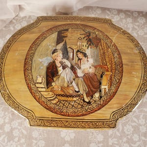 May include: An oval-shaped wooden tray featuring a central hand-painted illustration of two figures in traditional attire seated indoors. The border is decorated with intricate dark brown patterns on a light tan wood grain background.