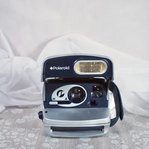 Peut inclure: Un appareil photo instantané Polaroid bleu marine et argenté. L'appareil photo est équipé d'un flash intégré et d'un objectif. Le logo Polaroid est visible sur le dessus. Conçu pour la photographie instantanée.