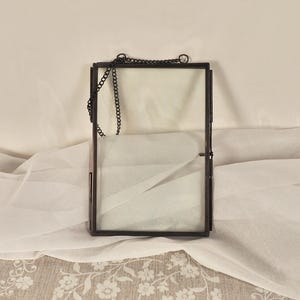 May include: A rectangular, bronze-colored glass frame with a chain for hanging. The frame has a hinged closure and is empty inside, with a white fabric background. The frame is designed to hold a photograph or other memento.