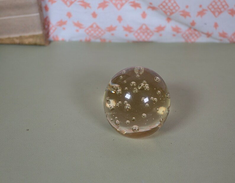 K&ouml;nnte beinhalten: Eine klare Glaskugel mit kleinen Luftblasen. Die Kugel liegt auf einer hellgrauen Oberfl&auml;che.