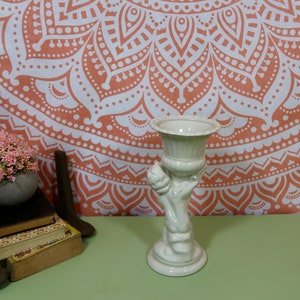 K&ouml;nnte beinhalten: Wei&szlig;er Keramik-Pflanztopf in Form einer Hand, die eine Vase h&auml;lt. Der Topf hat eine glatte, gl&auml;nzende Oberfl&auml;che und ist mit einem erhabenen Design verziert. Der Topf steht auf einer gr&uuml;nen Oberfl&auml;che.