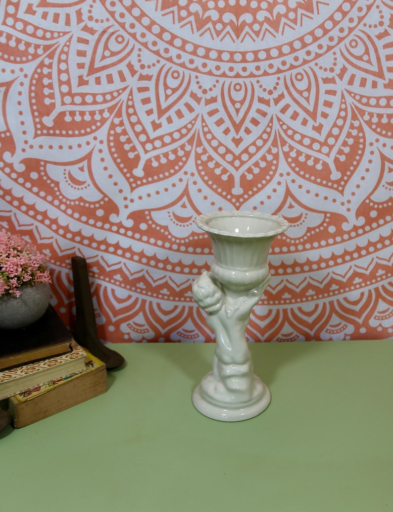 K&ouml;nnte beinhalten: Wei&szlig;er Keramik-Pflanztopf in Form einer Hand, die eine Vase h&auml;lt. Der Topf hat eine glatte, gl&auml;nzende Oberfl&auml;che und ist mit einem erhabenen Design verziert. Der Topf steht auf einer gr&uuml;nen Oberfl&auml;che.