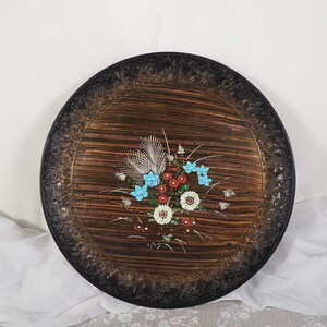 May include: Round decorative tray with a dark brown wood grain pattern and a black ornate border. The tray features a floral design with blue, red, and white flowers. A vintage-style serving piece.