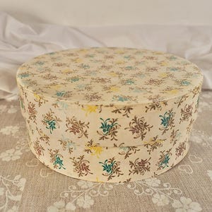 May include: A round, lidded storage box featuring a cream-colored base with a repeating floral pattern in shades of brown, teal, and yellow. The cylindrical container has a flat top and vertical sides, showcasing a consistent decorative print.