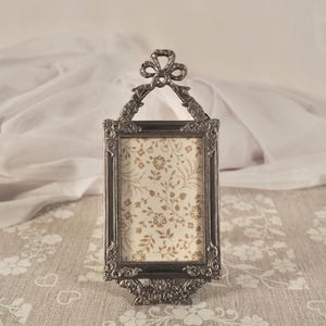 May include: An ornate, rectangular, silver-toned picture frame with a bow at the top and floral detailing. The frame holds a cream-colored floral patterned insert. The frame is approximately 13 cm tall.