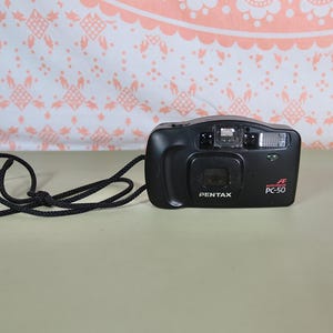 Puede incluir: Cámara compacta negra Pentax PC-50 con correa para el cuello negra. La cámara tiene forma rectangular con lente, flash y la marca "PENTAX" impresa en la parte delantera. La cámara está sobre una superficie verde claro.