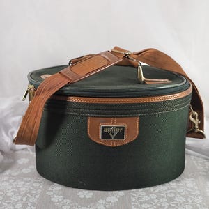 Può includere: Una borsa da viaggio cilindrica verde scuro con finiture in pelle marrone e tracolla. La borsa presenta una cerniera dorata e una toppa in pelle marrone con il marchio "antler". Progettata per trasportare oggetti da viaggio.