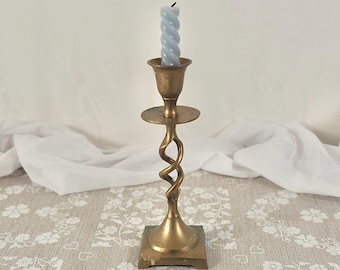 Può includere: Un candelabro in ottone con stelo attorcigliato e base quadrata contiene una candela blu chiaro a forma di spirale. Il candelabro ha una piccola piastra rotonda sotto il portacandele. Lo sfondo è un panno bianco e una tovaglia a motivi floreali.
