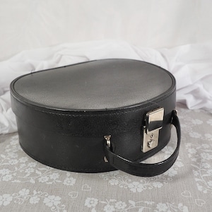 May include: A black, oval hat box with a silver clasp and handle. The box is made of a textured material and has a rounded top. The handle is attached to the side of the box with small metal hardware.