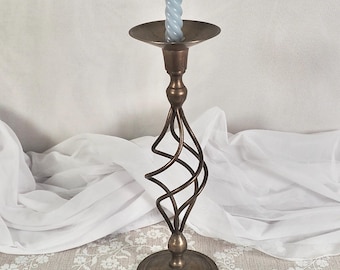 Può includere: Un candelabro in metallo color bronzo con un design a spirale sullo stelo, che tiene una candela attorcigliata blu chiaro. Il candelabro ha una base rotonda e una parte superiore circolare. Lo sfondo è un tessuto bianco.