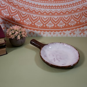 May include: A white ceramic dish with a brown rim and a brown handle. The dish is on a light green surface.