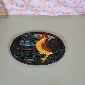 May include: Cast iron trivet featuring a rooster design. The rooster is yellow, red and black. The trivet is oval shaped and has a green and brown background.