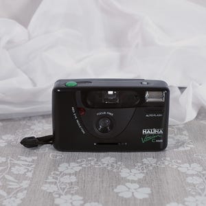 May include: Black Halina Vision C-XMS 35mm film camera with a green button and a wrist strap. The camera has "Focus Free" and "Auto Flash" features. The camera is on a floral patterned surface.