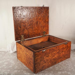 May include: A wooden box with an open lid, revealing the interior. The box is constructed from a dark brown, textured material, possibly particle board or plywood. The lid is attached with brass-coloured hinges and a latch. The box is designed for storage.