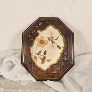 Puede incluir: Un marco de madera octogonal con acabado marrón oscuro, con un interior color crema con flores prensadas. El marco tiene detalles de hojas decorativas y un borde negro, creando una estética vintage.
