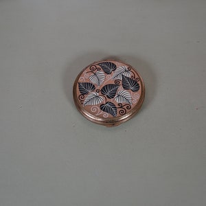 May include: A round, gold-coloured compact with a pink background and a black and white leaf design.