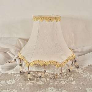 May include: Cream-coloured lampshade with a floral pattern. The shade is trimmed with gold-coloured lace at the top and bottom. Decorative beaded fringe hangs from the bottom edge. The lampshade is designed for a vintage or classic aesthetic, suitable for a living room.