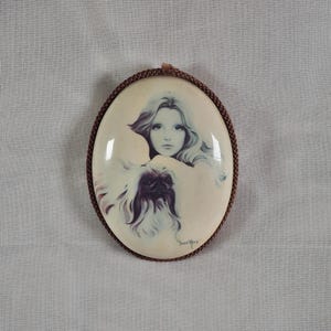 Puede incluir: Colgante ovalado con un retrato de una mujer de pelo largo y un perro pequinés. La obra de arte está en tonos de blanco, gris y marrón, con un borde de cuerda marrón. Firmado "David Young".