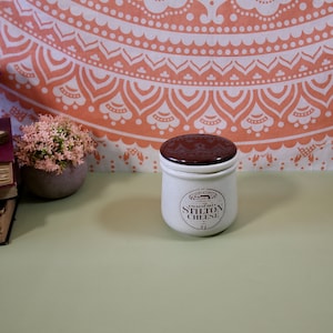 May include: A white ceramic jar with a brown lid. The jar has a label that reads "English Blue Stilton Cheese".