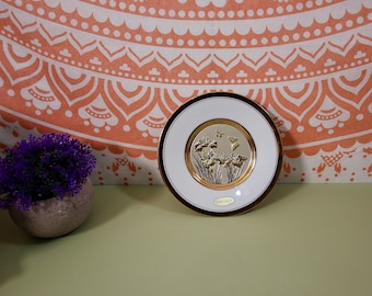 Könnte beinhalten: Ein weißer Keramikteller mit goldfarbenem Rand und einem erhabenen Design aus Blumen und Schmetterlingen. Der Teller trägt die Aufschrift "24kt Gold" auf der Unterseite.
