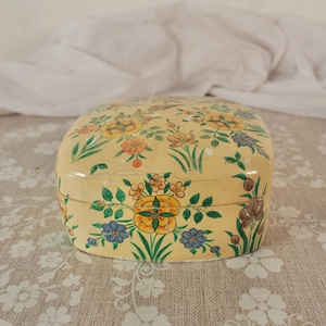 May include: A small, oval-shaped box with a cream-coloured base featuring hand-painted floral motifs in shades of yellow, orange, blue, and green. The surface has a glossy finish with visible fine cracks throughout the painted exterior.