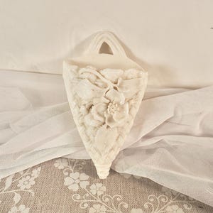 May include: A white ceramic wall pocket featuring a raised relief design of a central flower surrounded by leaves and a woven basket-weave texture. The piece has a triangular shape with a pointed bottom and a single arched opening at the top.