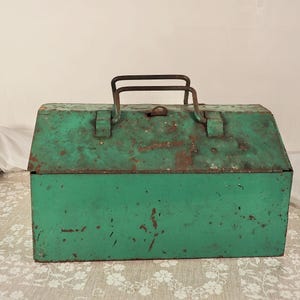 May include: A rectangular metal toolbox painted in a weathered green finish with visible rust spots throughout the surface. The box features a hinged lid with a central latch mechanism and a sturdy, dark-coloured metal handle attached to the top.