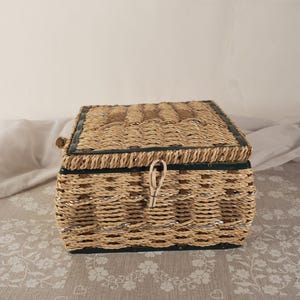 Puede incluir: Caja de almacenamiento rectangular tejida con tapa con bisagras. La caja está hecha de material tejido de color natural con un borde verde oscuro y un acento plateado. Tiene un cierre de lazo y palanca.
