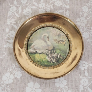 May include: A vintage gold-coloured decorative plate with a painted scene of a swan and cygnets on a grassy bank. The plate has a detailed, embossed border and a circular painting in the centre. The plate is round and has a raised edge.