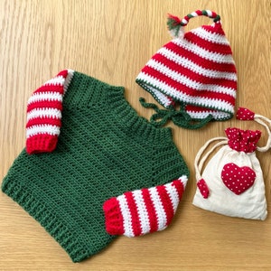 May include: A green crocheted sweater with red and white striped sleeves. The sweater has a round neckline and is made with a textured stitch. The sweater is paired with a red and white striped crocheted hat with a long white tassel.