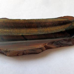 May include: A raw, rough-cut piece of tiger's eye gemstone. The stone is a deep brown color with gold and black stripes. It has a natural, irregular shape.