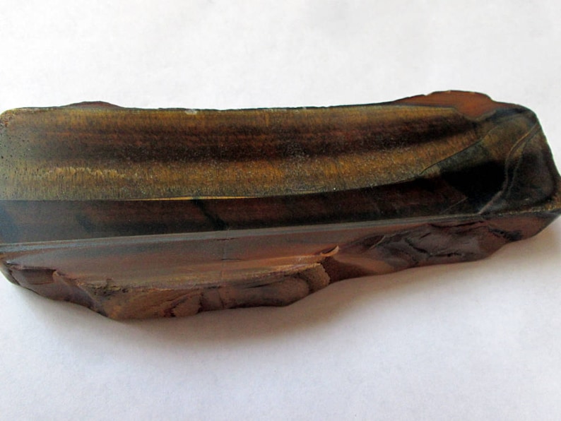 May include: A raw, rough-cut piece of tiger's eye gemstone. The stone is a deep brown color with gold and black stripes. It has a natural, irregular shape.