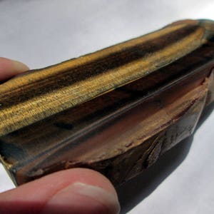 May include: A polished, rectangular piece of tiger's eye gemstone with a brown, gold, and black color pattern. The stone has a smooth, polished surface and a rough, natural edge.