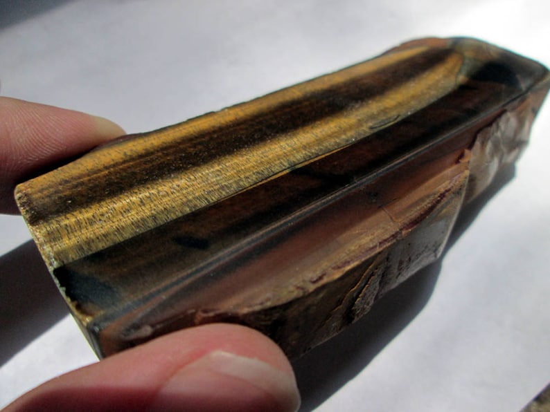 May include: A polished, rectangular piece of tiger's eye gemstone with a brown, gold, and black color pattern. The stone has a smooth, polished surface and a rough, natural edge.